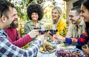 Happy friends having bbq dinner party in garden restaurant - Multiracial young people eating grill meat and drinking red wine in backyard - Food life style concept with guys and girls sitting outdoors
