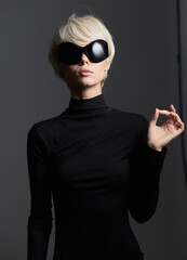 Stylish woman with short blonde hair wearing oversized black sunglasses and a form-fitting black turtleneck, posing confidently against a dark background.