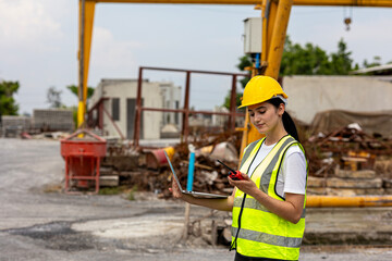 Attractive female engineer is using computer notebook and walkie talkie to communicate with her coworkers checking the construction work in a factory. Female technician with notebook.