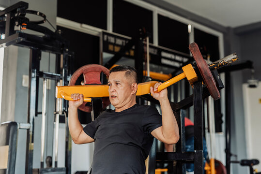 A fit, determined Southeast Asian man in his 50s performs a shoulder press on a plate-loaded machine at an open-air gym.
