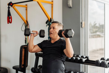 Southeast Asian man in his mid 50s performs an overhead dumbbell shoulder press at an open-air gym with proper form and dedication.