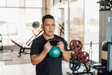 A focused Southeast Asian man in his 50s performs a goblet squat using a green kettlebell at an open-air gym.