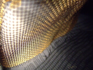 Close-up of a golden mesh fabric with metallic texture and intricate pattern, highlighted by dramatic lighting and shadows