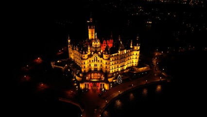 A night drone photo of Schwerin Castle in Northern Germany. The castle is beautifully lit and reflected in the surrounding lake. A peaceful, atmospheric image capturing historic architecture and winte