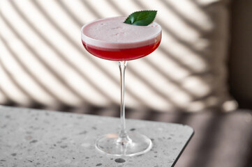 Elegant red cocktail in a coupe glass with a frothy top and a green leaf garnish, placed on a...