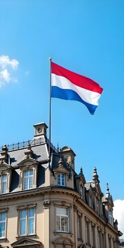 The Dutch flag waving on top of a historic building with ornate architecture, under a bright blue sky with a few clouds. The red, white, and blue stripes stand out against the elegant structure