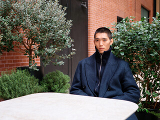 Portrait of handsome Chinese young man wearing black overcoat sitting in the street, young guy with black short hair all in black with urban background. Male fashion, cool Asian young man lifestyle.