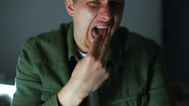 A guy in a dark room puts his fingers in his mouth to trigger a gag reflex.
The man feels sick and burps.
Food poisoning and nausea in a man.
Fingers in mouth