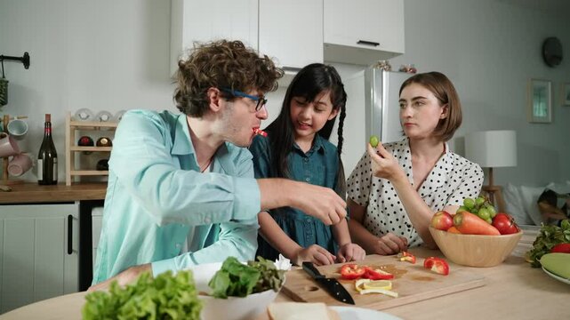 Attractive caucasian father and mother spend time together to cook fresh meal with asian daughter. Dad and mom preparing fresh vegetable for making salad at kitchen and eating vegetable. Pedagogy.