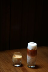 Two stylish drinks are served on a wooden table against a dark background—an iced layered latte with foamy top in a tall glass, and a creamy espresso or cortado in a short glass, cool and inviting.