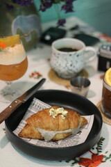 Croissant filled with cream and topped with edible gold leaf, served in a black dish. Surrounding it are a foamy cocktail, coffee, and a citrus drink.
