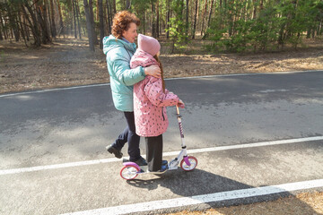 Grandmother teaches granddaughter to ride a scooter in the forest on a sunny day