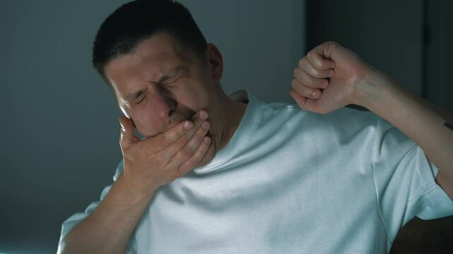 1111A young guy in a light T-shirt in the room yawns and rubs his face with his hands.
A man suffers from insomnia and cannot sleep at night.
Sleep problems.
Stress and overwork at work