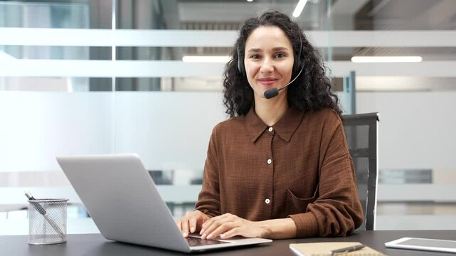 Portrait of young happy female corporate operator in headset sitting at desk at a workplace working on computer in modern business office. Smiling female agent of call center posing looking at camera