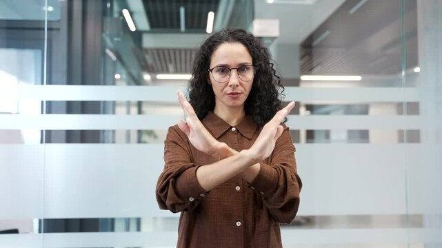 Serious businesswoman showing crossed arms, X sign, shaking head NO while standing in a modern business office looking at camera. The woman demonstrates a gesture of prohibition, denial, disagreement