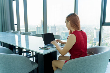 Young Asian woman in red dress working on laptop at modern high-rise office with panoramic city view, professional remote work and digital lifestyle concept © Piches