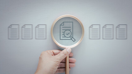 A hand holds a magnifying glass over a document icon that features a checkmark, suggests verification, approval, inspection, or selection over grey wooden background..