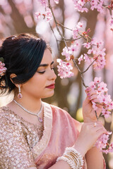 A woman in a shimmering pink traditional outfit stands serenely among cherry blossoms, radiating warmth, elegance, and cultural pride.