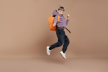 Cute schoolboy in headphones with backpack jumping on brown background