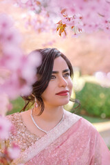 A woman in a shimmering pink traditional outfit stands serenely among cherry blossoms, radiating warmth, elegance, and cultural pride.