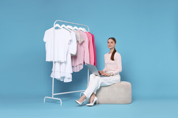Smiling fashion stylist with laptop and clothing rack on light blue background