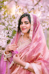 A woman in a shimmering pink traditional outfit stands serenely among cherry blossoms, radiating warmth, elegance, and cultural pride.