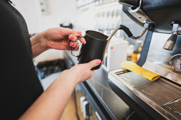 A barista steams milk using an espresso machine’s steam wand, holding a black metal pitcher. Steam rises as the milk froths, with a pressure gauge and a yellow cloth visible on the drip tray