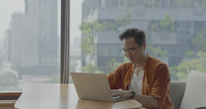 Senior Asian businessman wearing headset celebrates success during online meeting in modern co-working space. Remote work and business communication concept.