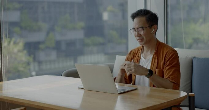 Senior Asian businessman wearing headset celebrates success during online meeting in modern co-working space. Remote work and business communication concept.
