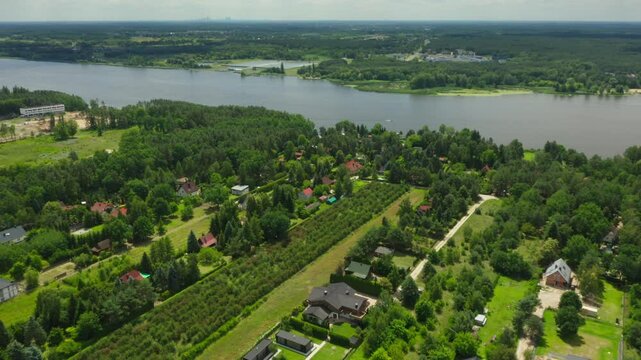 Approaching aerial view of Jachranka, a Polish village in Serock by the Narew River. The scene includes houses, private gardens, tree-lined areas, and the broad river, with Warsaw distant.