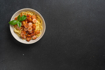 Spaghetti with sliced sausages and basil in white bowl on black background