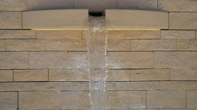 Jet d&rsquo;eau clair tombant d&rsquo;un bec encastr&eacute; dans un mur en pierre beige, vue frontale en plein jour, ambiance zen et rafra&icirc;chissante, design architectural moderne et minimaliste
