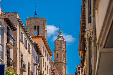 Church towers rise above traditional buildings on a quiet street in the old town of Alcañiz,...