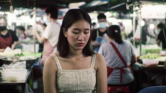 A young woman sits pensively among the lively atmosphere of a crowded night market. Vendors are busy with customers while colorful goods are displayed, creating an engaging environment.