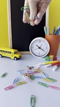 A hand holding colorful paper clips above a scattered arrangement of school supplies including a clock, a toy school bus, and a container with pens and dice on a bright yellow background.