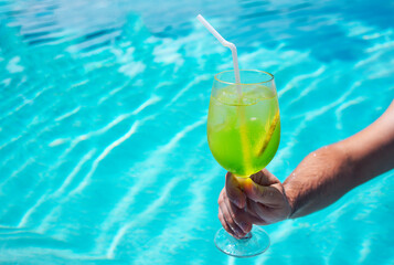 A delicious green cocktail in a glass in a male hand in the pool. A man holds a cup of cool summer drink with a straw and a slice of lemon.