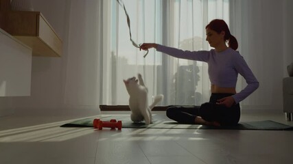 A woman sitting on a fitness mat engages her cat by using a toy on a rope in a living room. They share joyful, energetic playtime while promoting a healthy lifestyle. - Powered by Adobe