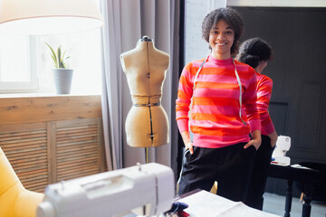 Young laughing African woman stands next to dressmaker mannequin. Smiling seamstress in casual clothes with tape measure. Charming black girl works in home workshop.