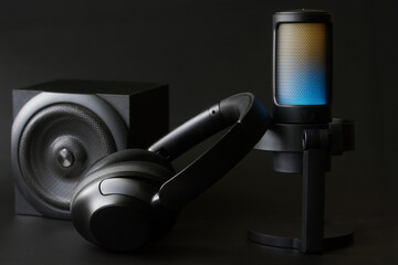A close-up of a modern condenser microphone with LED backlighting, over-ear headphones and speaker on a dark surface, perfect for showcasing podcast recording and live streaming setups. Photo
