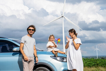 Concept of progressive happy family holding windmill toy and relax at wind farm with electric vehicle, happy family in the community with wind generators turbines, ev car