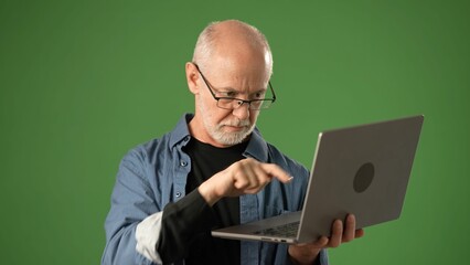 Senior man with glasses focuses on using a laptop, showcasing emotion and engagement against a solid green screen background, ideal for announcements.