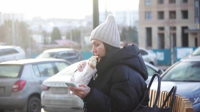 Woman sits on a bench in a park, savouring freshly baked bread while dressed warmly in winter attire