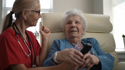 A caregiver helps an elderly woman operate a remote control while seated in a comfortable armchair. Natural light fills the cozy room, creating a warm atmosphere.