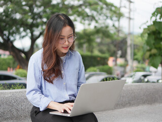 Young woman in blue striped shirt focused on her laptop while sitting on a stone bench in a leafy outdoor area.