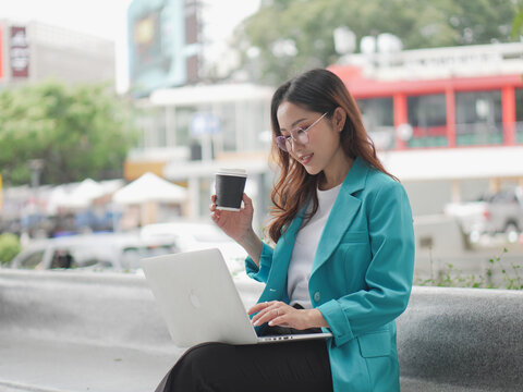 Woman in a teal blazer using a laptop outdoors while holding a coffee cup, with a soft smile and a relaxed posture.