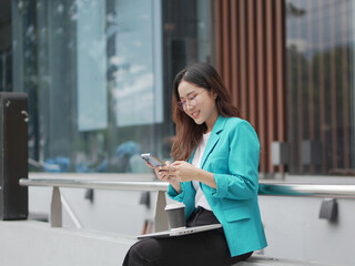 A smiling woman in a teal blazer is texting on her phone while sitting outdoors