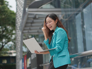 A cheerful woman in a teal blazer is holding a laptop and raising her fist in celebration outside