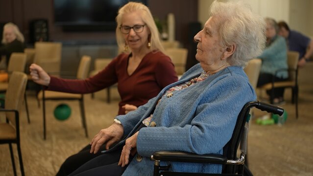 A lively atmosphere fills the community center as seniors participate in group activities. One smiling facilitator interacts with a resident in a wheelchair.
