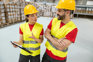 Female warehouse worker holding tablet, explaining tasks to male colleague with crossed arms.