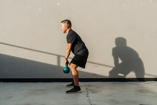 Fit middle-aged man outdoors holding kettlebell between legs, preparing for a swing exercise under late afternoon sunlight.